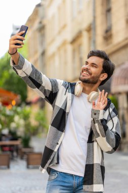 Neşeli Hintli blog yazarı akıllı telefondan selfie çekiyor, aile dostlarıyla internet üzerinden video görüşmesi yapıyor sosyal medya vlog 'u için hikayeler kaydediyor, el sallıyor. Şehir sokağındaki İspanyol adam..