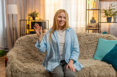 Genç bir kadın evinde oturuyor, sıcak dalgalar ve kolay iletişim için parlak bir gülümseme sunuyor. Kanepede beyaz bir kız arkadaşça el sallıyor merhaba, merhaba, günbatımı ışığı altında hoş geldin konuşması için ortam yaratıyor..