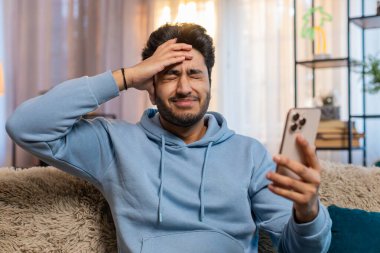 Akıllı telefon kullanan, negatif mesaj okuyan, oturma odasında kanepede oturmaktan rahatsız olan üzgün genç adam şok oldu. Alet sorunu olan Hintli adam üzgün görünüyor, telefonu tutuyor ve kötü habere tepki veriyor..