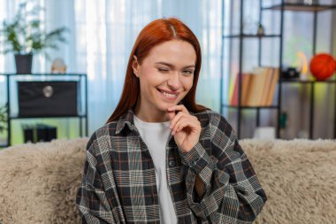 Genç bir kadın kameraya göz kırpıyor. Cilveli bir gülümsemeyle kendine güveni ve cazibesini gösteriyor. Kafkasyalı kızıl saçlı kız kanepenin üzerinde eğleniyor, duygularını ve kişisel çekiciliğini ifade ediyor.