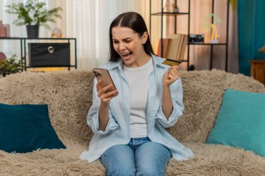 Evde genç bir kadın, engellenmiş hesap ya da hatalardan endişelenen akıllı telefondan kredi kartı bakiyesini endişeyle kontrol ediyor. Kanepedeki beyaz kız maddi sorunlar, kayıp ya da başarısız ödemeler konusunda vurguladı