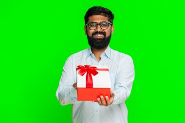 Hintli bir adam gülümseyerek hediye kutusunu tutarak elini uzatıyor ve birini cömert bir sürpriz olması için davet ediyor. Yeşil renkli arka planda izole edilmiş bir Arap adam kibarlık göstererek mutlu bir jest yapıyor..