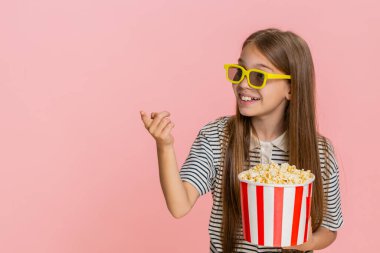 Genç kız mutlu bir şekilde patlamış mısır yiyor, televizyonda eğlenceli bir çizgi film izliyor, neşeli bir şekilde gülüyor, geniş bir kesime gülümsüyor, eğlenceli animasyon sahnelerinden haz alıyor. Pembe arka plandaki okul çocuğu.