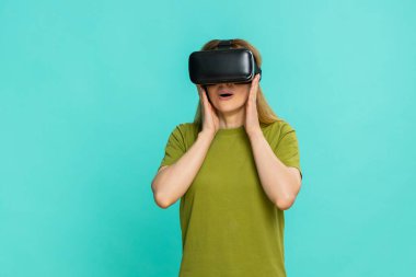 Orta yaşlı kadın VR kulaklık takıyor ve dikkatlice etrafa bakıyor, meraklı odaklı yüzüyle dijital çevreyi keşfediyor. Mavi arka planda izole edilmiş kız sanal dünyaya ilgi göstererek tepki veriyor