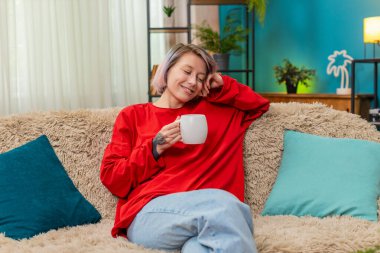 Evde sakince kahve içen genç bir kadın, rahatlamış, tatmin olmuş, huzurlu bir zevk ve rahatlık ifade ediyor. Mutlu kız sessizce gülümsüyor, rahatlıyor, kanepede içecekle keyifli vakit geçiriyor..