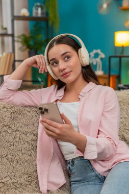 Genç bir kadın evdeki kanepede kablosuz kulaklık takarak oturuyor ve akıllı telefonuyla podcast dinliyor ve notlar yazıyor. Beyaz kız yeni bilgilerden memnun ve öğrenirken ritim tutuyor..