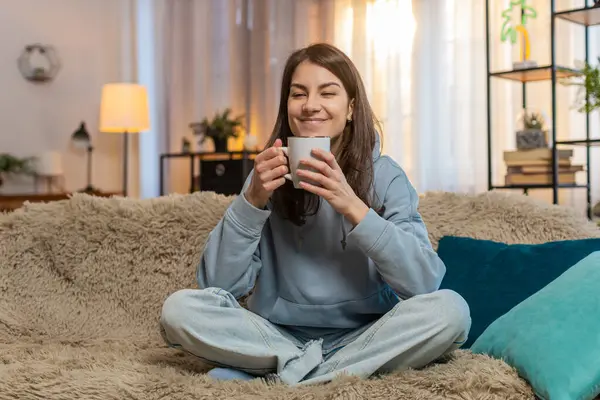 Mutlu genç bir kadın, içecek kokusu alıyor sıcak kahve içiyor, mola veriyor, dinleniyor. Gülümseyen beyaz kız aroma çayını tek başına içmeyi sever. Oturma odasındaki kanepede oturur..