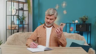 Beyaz, olgun bir adam evde oturuyor cep telefonu hoparlöründen konuşuyor, not defterine notlar yazıyor. Orta yaşlı adam odaklanmış, kararlı görünüyor, verileri analiz ediyor ve çözümler arıyor