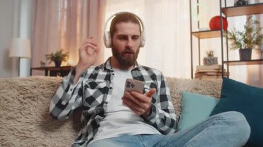 Genç adam kablosuz kulaklıklarla evde titrer müzik dinler, akılda kalıcı bir ritimle başını sallar. Kanepedeki beyaz adam başparmağını kaldırdı, bu da harika bir melodiyi onayladığını gösteriyor.