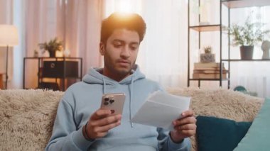 Uygulama yoluyla faturaları ödeyen genç Hintli adam elinde akıllı telefon ve makbuzlarla evdeki oturma odasındaki kanepede oturuyor. Arap adam doğru ödeme detaylarını özenle gözden geçiriyor.