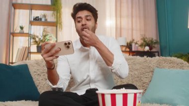 Hintli bir adam akıllı telefondan videoyu izlerken kanepede patlamış mısır yiyor. Dijital eğlenceye dalmış eğlencenin ve heyecanın tadını çıkaran Arap bir adam.