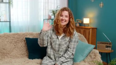 Evdeki genç kadın sıcak bir şekilde gülümsüyor ve sıcak ve neşeli bir şekilde merhaba demek için el sallıyor. Kanepede oturan beyaz saçlı kız neşe, hoş geldin jesti ve olumlu duygusal bağlarını dile getiriyor.