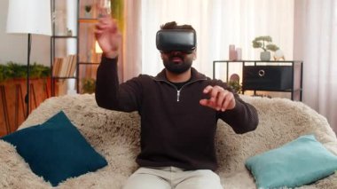 Evde oturan Hintli adam VR kulaklığı takıyor ve sanal dünyaya tepki göstererek şaşkınlıkla başını hareket ettiriyor. Arap adam heyecan ve enerji gösteriyor, tamamen fütüristik üç boyutlu dijital deneyime dalmış..