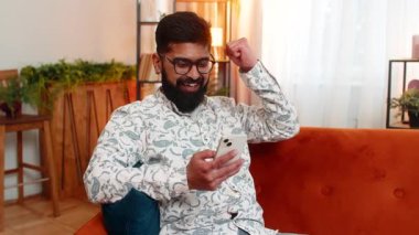 Evdeki Hintli adam akıllı telefondan gelen iyi haberi koltukta otururken ve heyecanlıyken kutluyor. Arap adam gülümseyerek yumruğunu kaldırıyor zafere ve başarıya olumlu tepki gösteriyor.