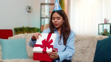 Hintli genç bir kadın doğum günü hediyesini kanepesinde açar ve şaşırmış bir ifadeyle ve büyük bir gülümsemeyle tepki verir. Arap kızı heyecan, neşe, mutluluk ve sıcak şaşkınlık gösteriyor..