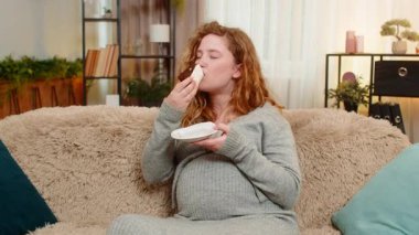 Hamile genç bir kadın, elinde bir parça pasta tutarak kanepesinde oturuyor, neşeli bir ifadeyle yemek yiyor, tatlı tadını çıkarıyor. Tasasız kız küçük zevkleri kucaklıyor, sade mutluluğu.