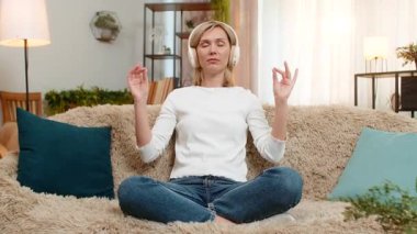 Orta yaşlı bir kadın, meditasyon pozunda, gözleri kapalı ve kulaklıkları açık bir şekilde kanepeye oturur. Yetişkin kadın sakinleştirici müzik dinliyor, huzurlu bir zihinsel durumda dinleniyor ve duygusal olarak yere çakılmış hissediyor..