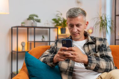 Evdeki oturma odasında cep telefonu tebessümleri kullanarak kanepede oturan beyaz bir genç. Gri saçlı mutlu adam mesajlaşırken akıllı telefon sosyal medya uygulamaları içeriğini paylaşıyor.