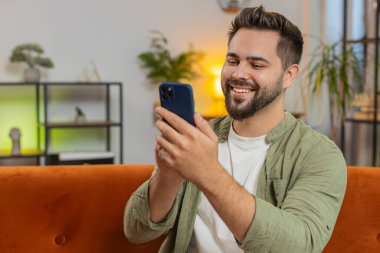 Evdeki oturma odasında gülümseyen cep telefonuyla kanepede oturan mutlu beyaz adam. İnternet üzerinden akıllı telefon sosyal medya uygulamalarında mesajlaşan adam film izliyor.
