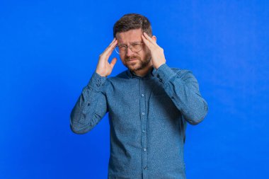 Keyifsiz, orta yaşlı, gözlüklü, baş ağrısı problemini tedavi etmek için şakak ovalayan orta yaşlı beyaz bir adam migren stresinden muzdarip. Acı içinde kıvranan bir adam, mavi arka planda yüksek tansiyon.