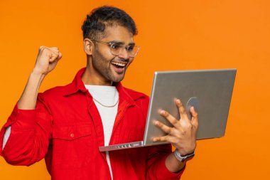 Heyecanlı mutlu Hintli adam dizüstü bilgisayarda yazıyor, proje üzerinde çalışıyor, sonuçların tadını çıkarıyor, piyango oyunu kazanıyor, başarıyı kutluyor, online alışveriş yapıyor, zafer kazanıyor. Turuncu arka planda izole edilmiş Arap adam.