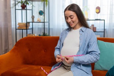 Kanepede gülümseyen hamile bir kadın. Ölçü bandıyla göbeğini ölçüyor. Doğumu bekleyen genç bir anne, vücut ölçülerini değiştiriyor. Hamilelik annelik kavramı.