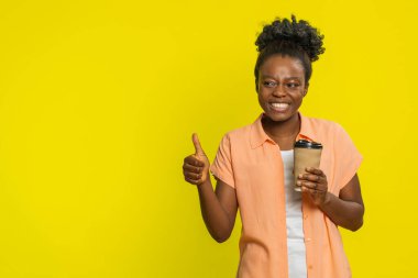 Afro-Amerikalı bir kadın elinde bir fincan plastik kahve tutuyor, aroması ve tadı ile mutlu ve tatmin olmuş bir bakışla. Sarı arka planda izole edilmiş siyah bir kız. Keyif verici. En sevilen sabah içme ritüelinden.