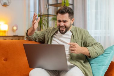 Tartışma. Öfkeli, sinirli, serbest çalışan genç adam koltukta oturmuş hararetli sohbetlere dalıyor. Kafkasyalı erkek, apartman odasındaki kanepede dizüstü bilgisayar kullanarak hayal kırıklığı yaşıyor..