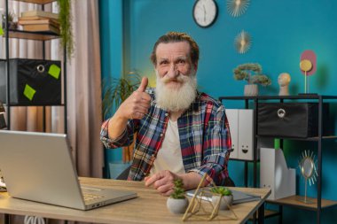 Gibi. Gülümseyen Kafkas kıdemli bir adam kameraya onaylayıcı bir şekilde bakıyor. İki başparmak işareti gösteriyor. İyi haber olumlu geri bildirim. Ev ofis masasında dizüstü bilgisayarı olan mutlu büyükbaba..