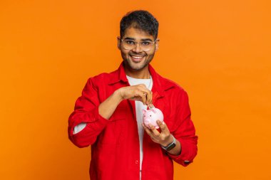 Mutlu Hintli adam gelecek ihtiyaçlar için kumbaraya dolar harçlığı koyuyor. Bozuk paraları olan Arap genç adam gelir artışını finanse ediyor. Maaş artışı, turuncu arka planda ekonomi.