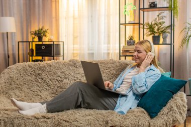 Genç bir kadın evde serbest çalışmaktan, dizüstü bilgisayarla yazmaktan ve kulaklıklarla sessiz müzik dinlemekten hoşlanıyor. Kanepede dinlenen kız, sıcak gün batımı, rahat, profesyonel ve yaratıcı bağlılık.