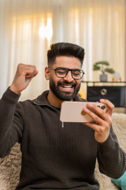 Evde Hintli bir adam akıllı telefona bakar ve iyi haberler aldıktan sonra büyük bir gülümseme ve yumruğunu sıkarak kutlar. Arap adam kanepede tek başına oturuyor, neşe ve heyecanı neşeli bir hareketle ifade ediyor.