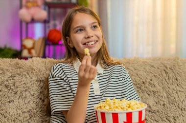 Genç kız patlamış mısır yer ve kanepede oturup eğlenirken televizyonda komik çizgi filmler izler. Okullu çocuk evde rahat bir film izlerken keyifli ve rahat görünüyor.