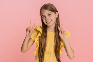 Genç kız kameraya sımsıkı gülümserken iki parmağıyla barış işareti yapıyor, neşe ve pozitiflik saçıyor. Pembe arka planda izole edilmiş, neşeli ve genç ruhlarla dolu bir okul çocuğu..