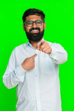 Hintli adam doğrudan kameraya bakar kendine güvenen bir gülümsemeyle, kişisel tercihini belirterek, sıranın sende olduğunu söyler. Yeşil renk arka planda izole edilmiş Arap adam karar ve seçim jestini gösteriyor