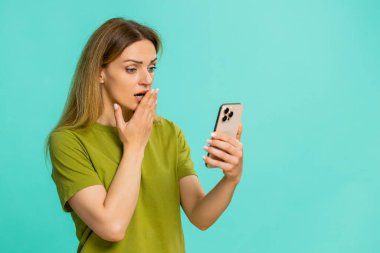 Orta yaşlı bir kadın akıllı telefon ekranına korkmuş görünüyor. Ani videoya ya da kötü habere tepki veriyormuş gibi ağzı şoka sokuyor. Mavi arka planda izole edilmiş bir kız korku, şaşkınlık ve duygusal gerilimi ifade eder.