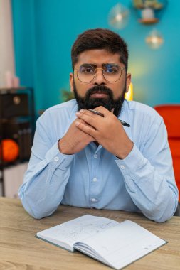 Evindeki Hintli adam kalemini elinde tutuyor. Dikkatlice ileriye bakıyor ve odaklanmış bir zihinle görevler planlıyor. Yakın plan Arap adam bir sonraki hamle ve stratejik fikirler üzerinden kalem çevirme hedefleri düşünüyor