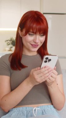 Evde genç bir kadın akıllı telefonu kontrol ediyor. İyi haber atlamaları başarıyı kutlar. Kanepede oturan kızıl saçlı kız yumruğunu kaldırıyor. Mutlu tepki veriyor.