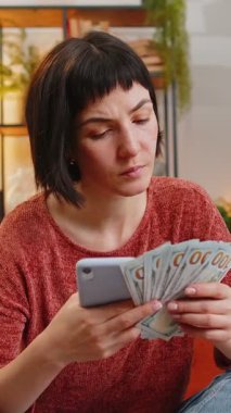 Gülümseyen genç bir kadın evdeki kanepede akıllı telefon hesap makinesi kullanarak para banknotlarını sayıyor. Hispanik mutlu kız daire için bütçe, tasarruf, kazanç ve planlama tatili hesaplıyor