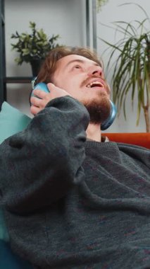 Kablosuz kulaklıklı mutlu, gevşemiş Kafkasyalı genç adam evinde kanepede dans ediyor ve akıllı telefon eğlencesinde favori disko müziğini dinliyor. İnsanlar hafta sonu aktiviteleri