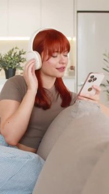 Genç ve mutlu bir kadın akıllı telefonuyla kulaklıkla müzik dinliyor ve vücut ve zihni rahatlatıyor. Kanepede gülümseyen kızıl saçlı kız. Sakin ol. Dinlen ve rahatla.