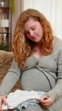 Hamile genç kadın, bebek kıyafetlerini evde koltukta otururken, küçük kıyafetleri nazik bir gülümsemeyle katlarken, sevgi ve beklenti hissederken sınıflandırıyor. Mutlu kız gelecekteki annelik ve aile bağlarına hazırlanıyor..
