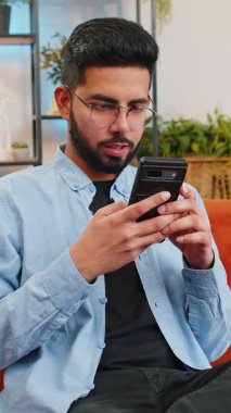 Gündelik giysiler içindeki mutlu genç adam akıllı telefondan düşünüyor ve mesajlaşıyor. Hintli adam kanepede oturuyor ve evdeki oturma odasında cep telefonu kullanıyor. Erkek tarayıcı internetinden bilgi arıyor.