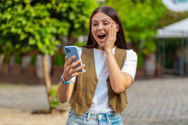 Aman Tanrım. Vay canına. Heyecanlı mutlu, neşeli Avrupalı genç kadın akıllı telefon daktilo kullanıyor kutlama yapıyor açık havada iyi haber kazan. Şehir caddesinde duran yetişkin kız. Kasaba yaşam tarzları