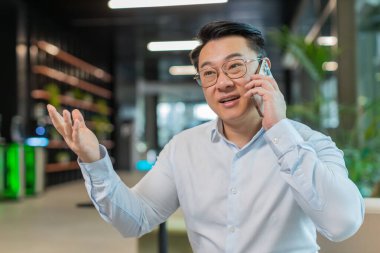 Tişörtlü genç işadamı cep telefonuyla müşteriye cevap veriyor, koltukta oturarak akıllı telefon kullanıyor. Sohbet. İnternette çalışan Çinli adam, modern ofis lobisinde konuşuyor..