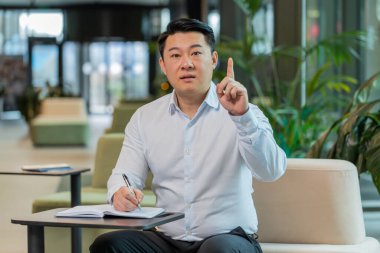 Olgun Çinli iş adamının, iş arkadaşının modern ofis lobisindeki görüntülü sohbete günlük yazarken konuşurken kameraya baktığı görüntüler. Yönetici masada notlar alıyor. Web kamerası görünümü.