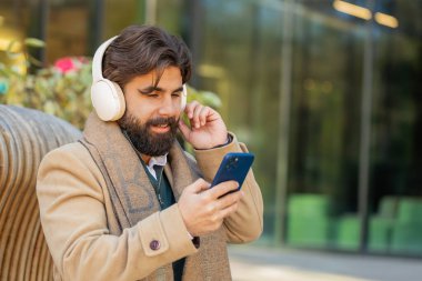 Kablosuz kulaklıkla dans eden mutlu, rahat Hintli genç işadamının portresi, akıllı telefon dansında enerjik disko müziği dinlemek. Lübnanlı sakallı adam şehir merkezindeki bankta oturuyor.