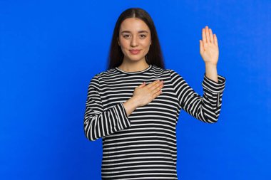 Dürüst olacağıma yemin ederim. İçten, sorumluluk sahibi genç bir kadın yemin etmek için elini kaldırıyor, dürüst olacağına ve doğruyu söyleyeceğine söz veriyor, elini göğsüne koyuyor. Mavi stüdyo arka planında izole edilmiş bir kız. Yaşam Tarzları