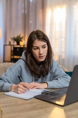 Genç, beyaz bir kadın dizüstü bilgisayar kursunu izliyor kanepenin üzerinde oturuyor ve not defterine not alıyor. Düşünceli kadın serbest çalışan, günlük giysiler içinde evden oturma odasına çalışıyor..