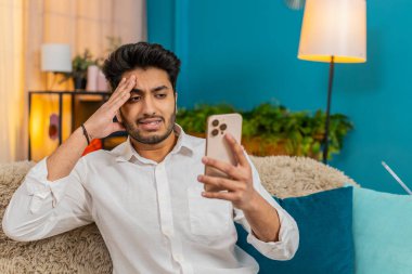 Evde oturan Hintli adam üzgün bir şekilde alnından akıllı telefon eline bakarak kötü haberler okuyor. Stresli hissediyor. Adam olumsuz mesaja tepki veriyor, endişeli, şok olmuş, beklenmedik bir bilgiyi işliyor.
